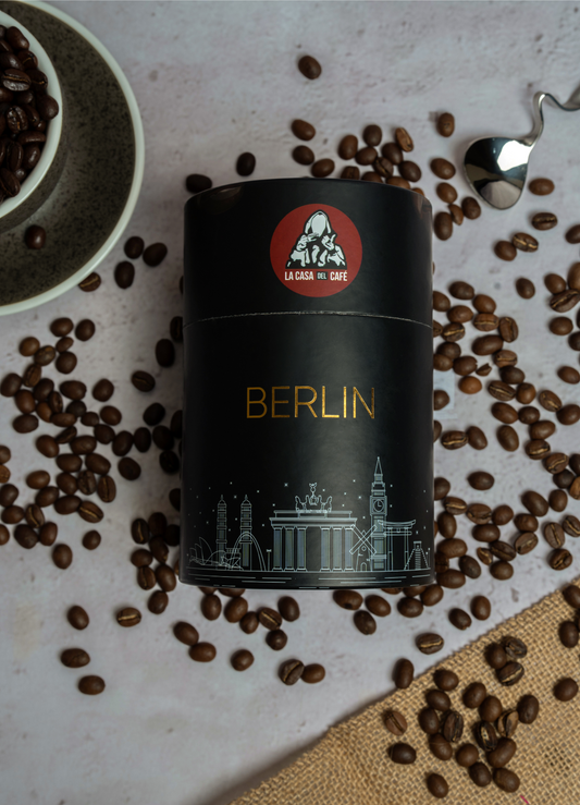 A photograph of a coffee tin labeled 'Berlin' from 'La Casa del Café' surrounded by scattered coffee beans, with iconic Berlin landmarks illustrated in white on the tin. The image also features a ceramic bowl filled with coffee beans and a metal spoon on a textured surface