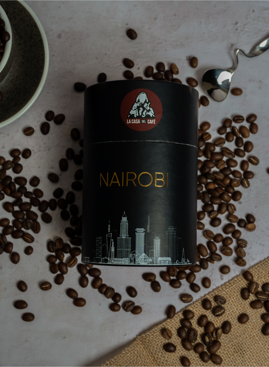 Image of a coffee can labeled 'NAIROBI' from 'La Casa del Café'. The can is black with a red circular logo featuring a hooded man giving a thumbs up. Below the logo, the can is adorned with a white line drawing of Nairobi's skyline, including skyscrapers and iconic buildings, highlighted with gold accents. The can is surrounded by scattered coffee beans, a ceramic bowl partially filled with more beans, and a silver spoon on a textured surface