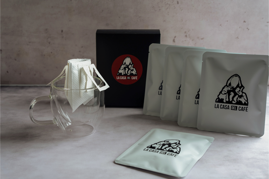 "Image showing coffee products from 'La Casa del Café'. The scene includes a transparent glass coffee mug with a unique, flat design and several white, flat coffee filter bags hanging over its edge. Next to the mug, there is a black box with a red and white logo of a hooded man giving a thumbs up. To the right, several matte white coffee packets stand upright, each featuring the same logo in black. The setup is displayed against a textured, neutral-colored background."