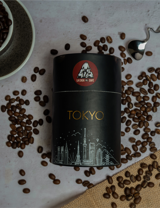 Image of a coffee can labeled 'TOKYO' from 'La Casa del Café'. The can is black with a red circular logo featuring a hooded man giving a thumbs up. Below the logo, the can is adorned with a white line drawing of Tokyo's skyline, including landmarks like the Tokyo Tower and modern skyscrapers, highlighted with gold accents. The can is surrounded by scattered coffee beans, a ceramic bowl partially filled with more beans, and a silver spoon on a textured surface.